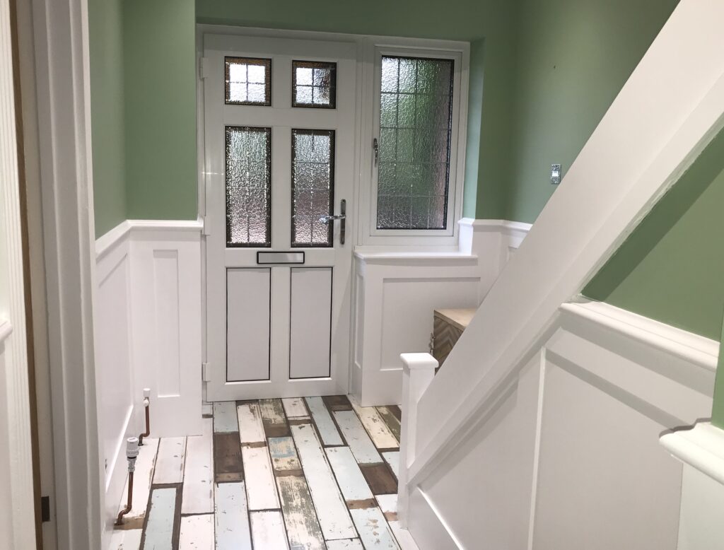 Picture depicts a stylish hallway with shaker wall panelling the panels are painted white and the wall above is painted Green